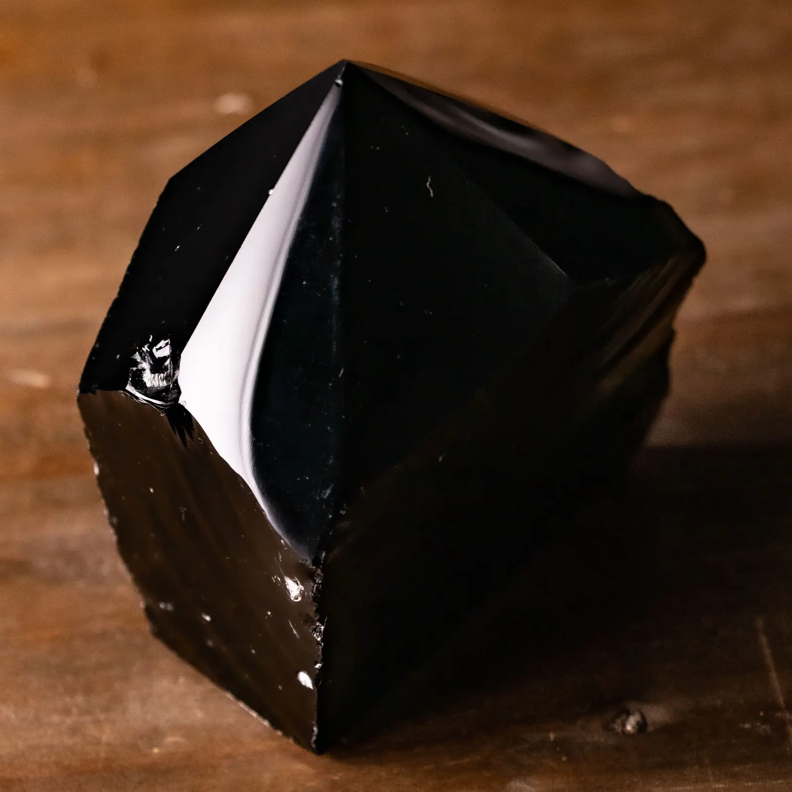 A polished obsidian point on warm timber against black, its faces breaking to a glassy edge